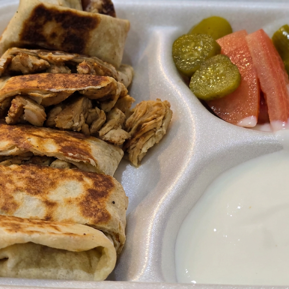 Grilled chicken wraps served in a compartmentalized takeout container with pickles, tomato slices, and creamy white sauce.