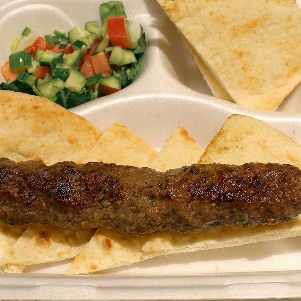 Grilled kofta kebab served over toasted flatbread with pita wedges and chopped cucumber-tomato salad in a divided tray.